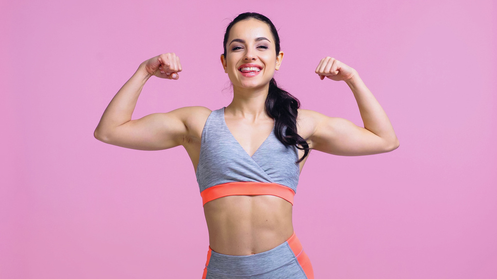 vrolijke sportvrouw tonen spieren geïsoleerd op roze - Foto, afbeelding