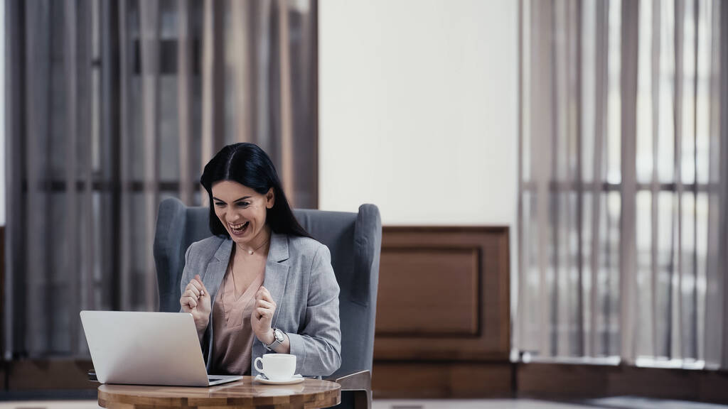 возбужденный фрилансер смотрит на ноутбук возле чашки кофе на столе в вестибюле ресторана - Фото, изображение