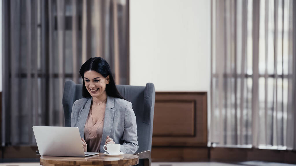 happy woman looking at laptop near cup of coffee on table in lobby of restaurant - Foto, afbeelding