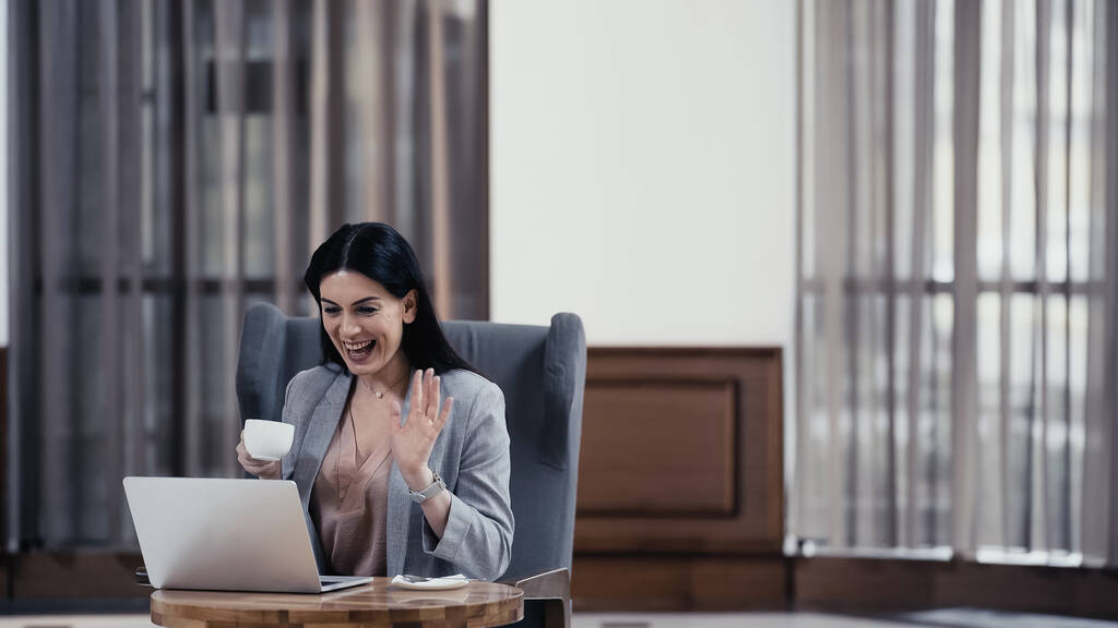 glückliche Frau mit Pokal und winkender Hand bei Videoanruf am Laptop  - Foto, Bild