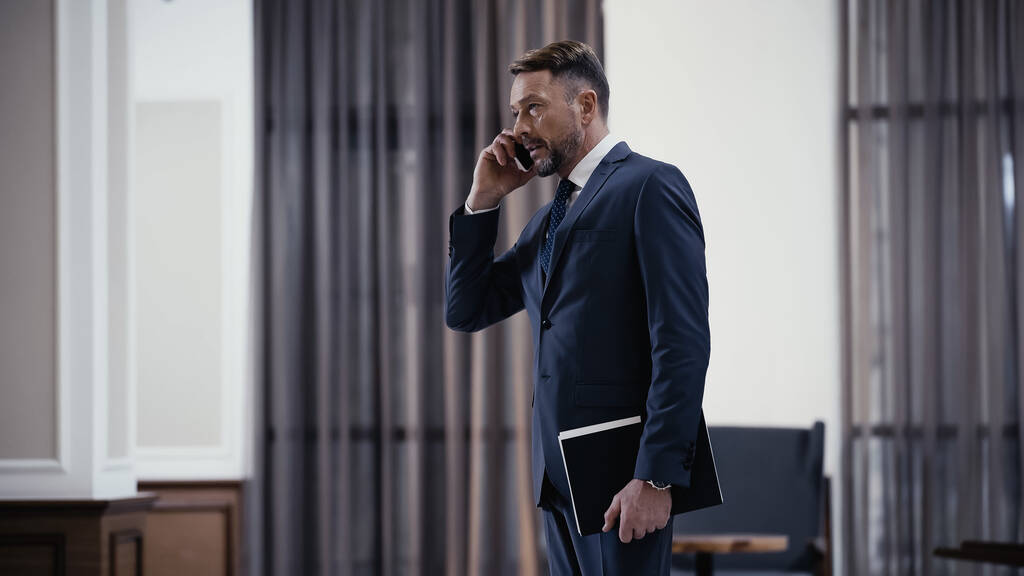 bearded businessman in suit talking on smartphone and holding paper folder in restaurant  - Fotografie, Obrázek