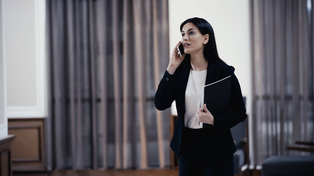 Businesswoman with paper folder talking on smartphone in lobby of restaurant - Φωτογραφία, εικόνα