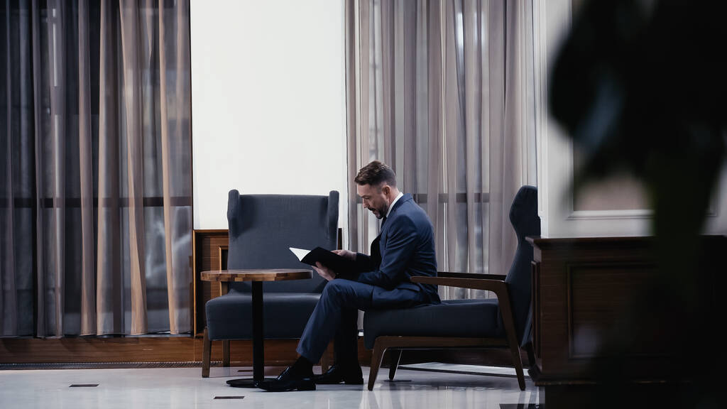 Geschäftsmann im Anzug sitzt auf Sessel und schaut sich Dokumente an  - Foto, Bild