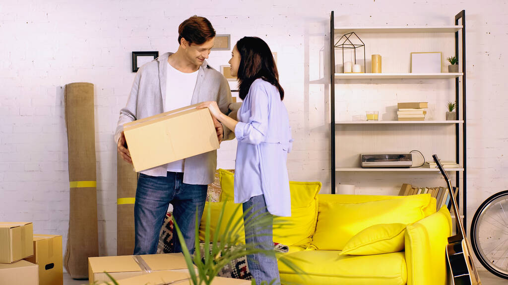 happy young man holding box near cheerful girlfriend in living room - Photo, Image