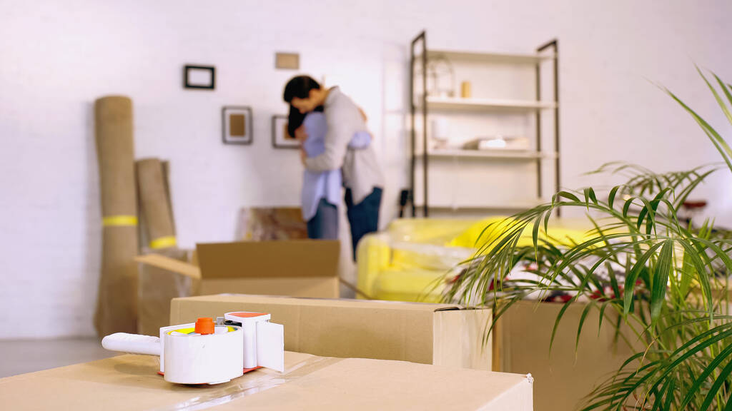 cinta adhesiva en cajas cerca de borrosa pareja abrazándose en un nuevo hogar - Foto, imagen