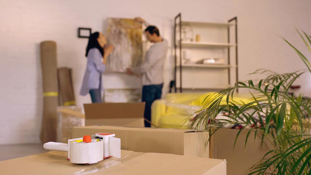 cinta adhesiva en cajas de cartón cerca de pareja borrosa colgando pintura en un nuevo hogar - Foto, imagen