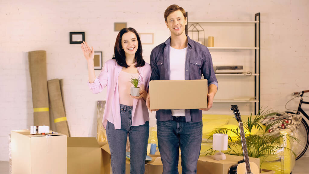 šťastná žena mává rukou a drží rostlina v blízkosti muže s krabicí v novém domově - Fotografie, Obrázek