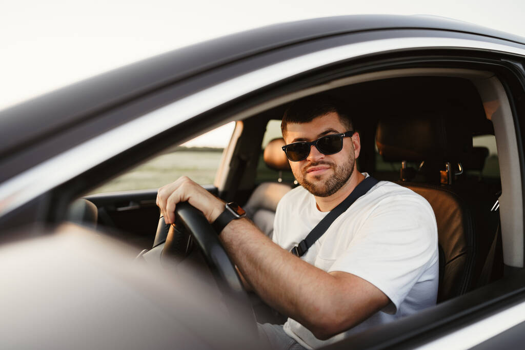 Szakállas férfi napszemüvegben ül a kocsijában és a kamerába néz. - Fotó, kép