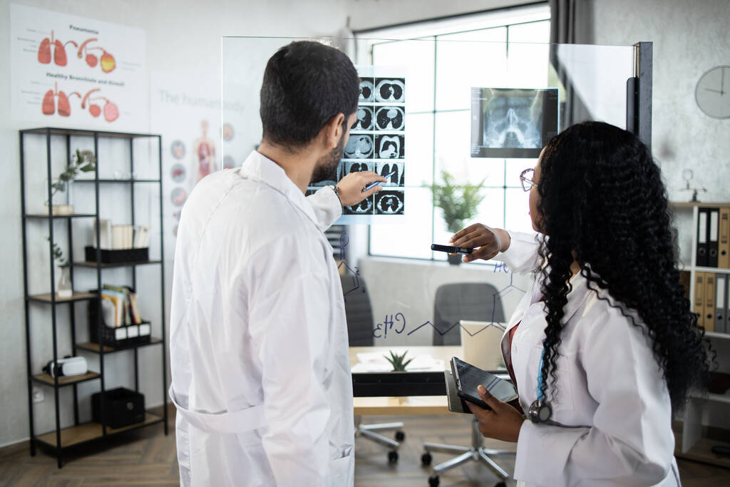 Kaksi pätevää monikansallista lääkäriä, jotka seisovat toimiston lasilevyn lähellä ja tulkitsevat CT-skannausta - Valokuva, kuva