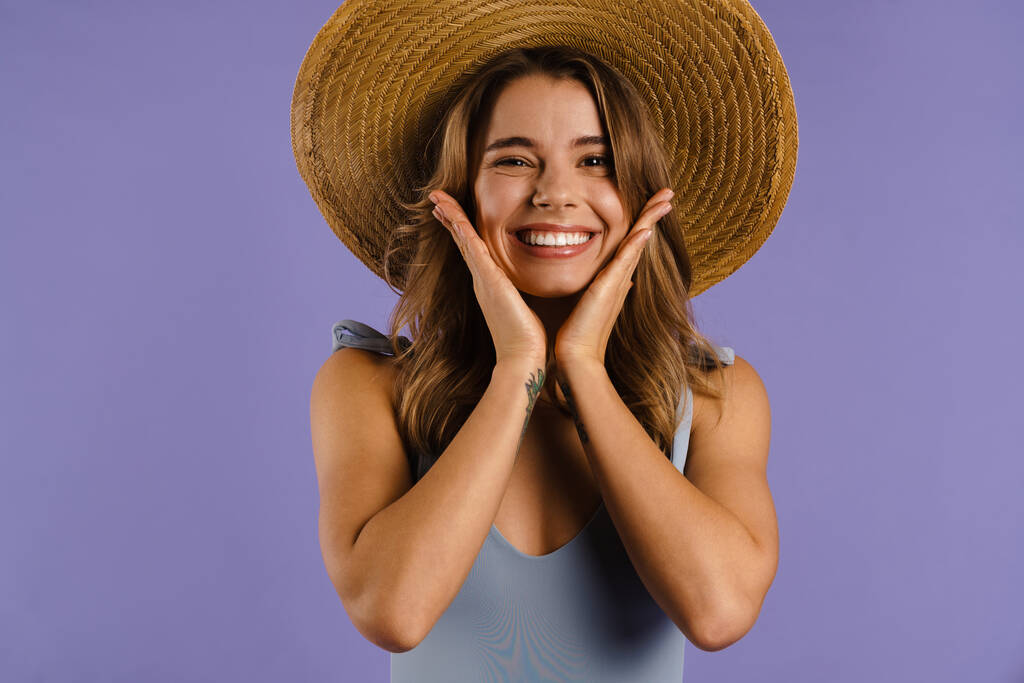 Молода біла жінка в солом'яному капелюсі посміхається і схоплює її обличчя ізольовано на фіолетовому фоні
 - Фото, зображення