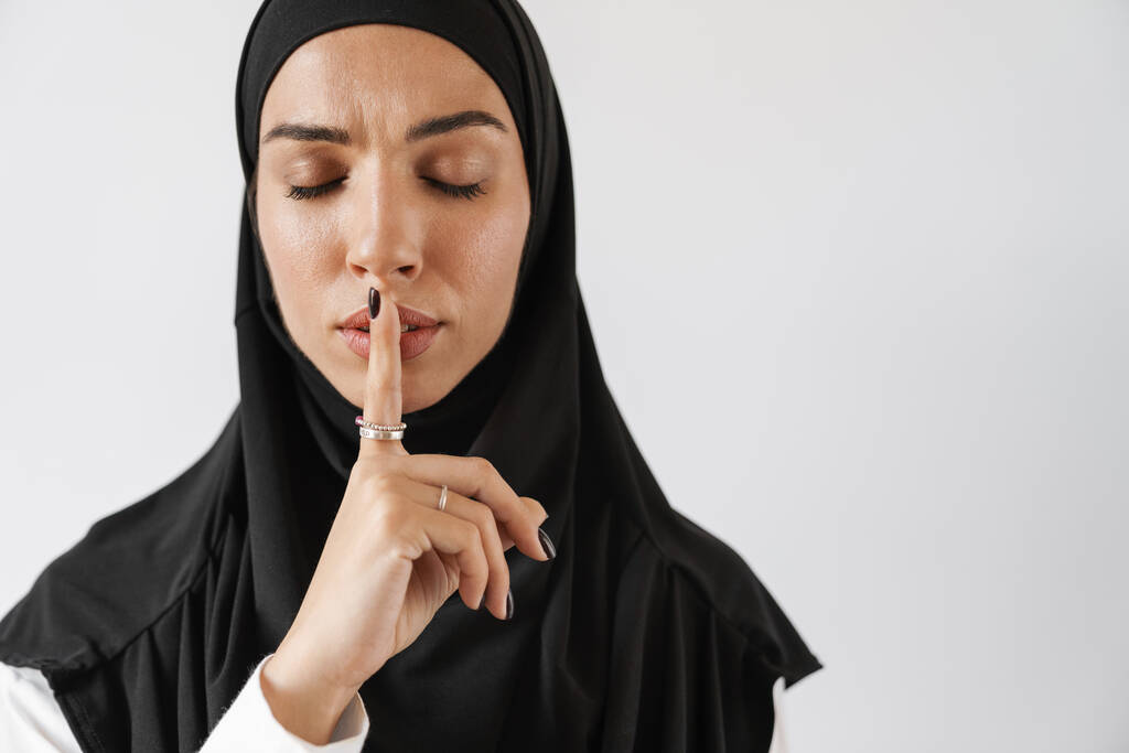 Ένα πορτραίτο της γυναίκας με αυτοπεποίθηση μουσουλμάνα στο χιτζάμπ δείχνει μια χειρονομία σιωπής με κλειστά μάτια ενώ στέκεται στο λευκό στούντιο - Φωτογραφία, εικόνα