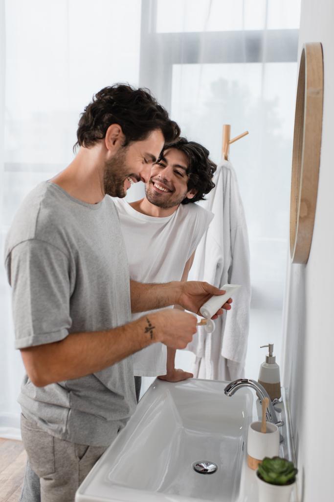 Χαρούμενος γκέι άντρας που κοιτάζει τον φίλο του να πιέζει οδοντόκρεμα στο μπάνιο  - Φωτογραφία, εικόνα