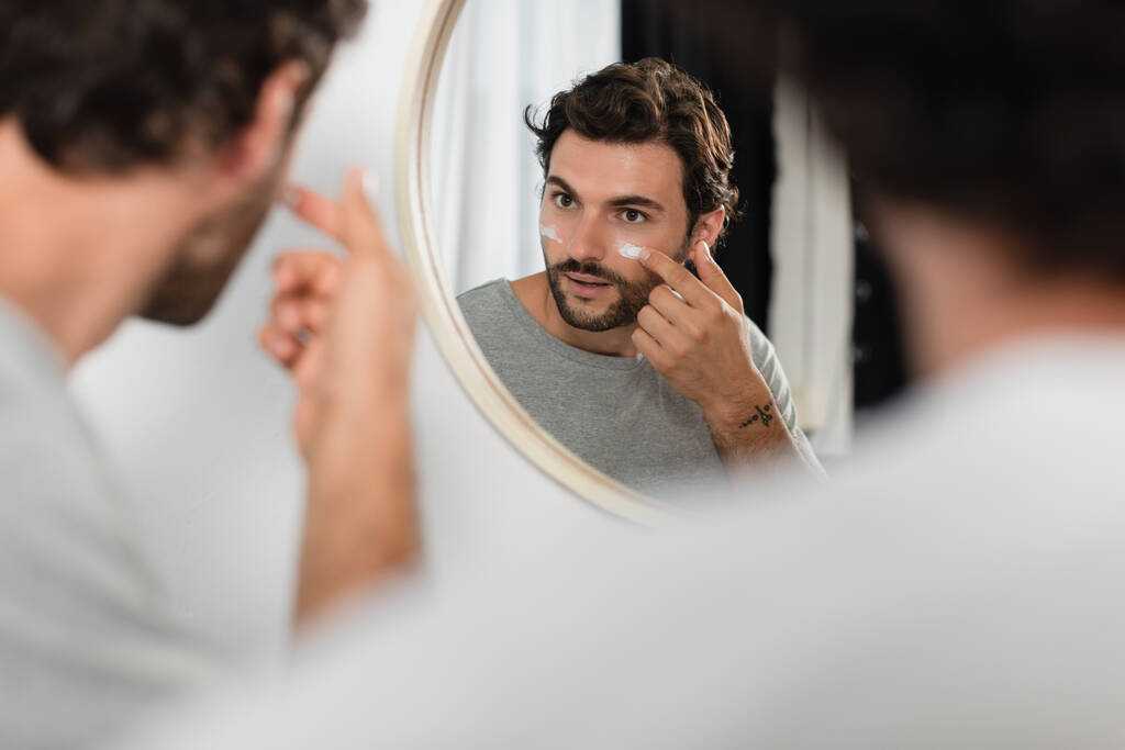getatoeëerde man aanbrengen crème op gezicht in de buurt wazig vriendje in de badkamer - Foto, afbeelding