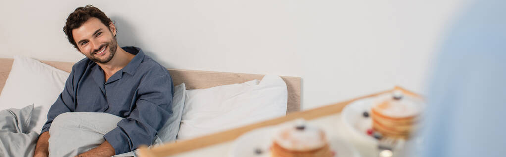 hämärtynyt mies tilalla aamiainen tarjotin pannukakkuja lähellä onnellinen poikaystävä sängyssä, banneri - Valokuva, kuva