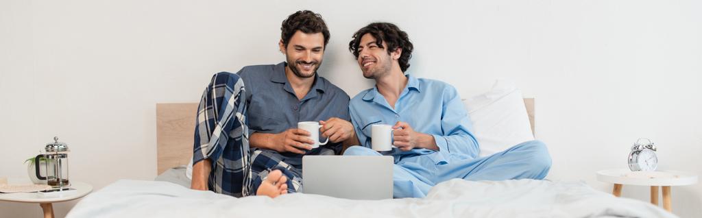счастливые геи смотрят кино на ноутбуке и держа чашки чая, баннер - Фото, изображение