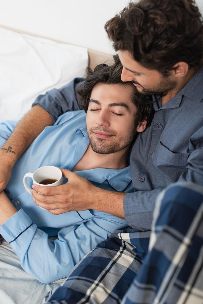 入れ墨ゲイの男保持カップのお茶と巨大ボーイフレンドでベッド - 写真・画像