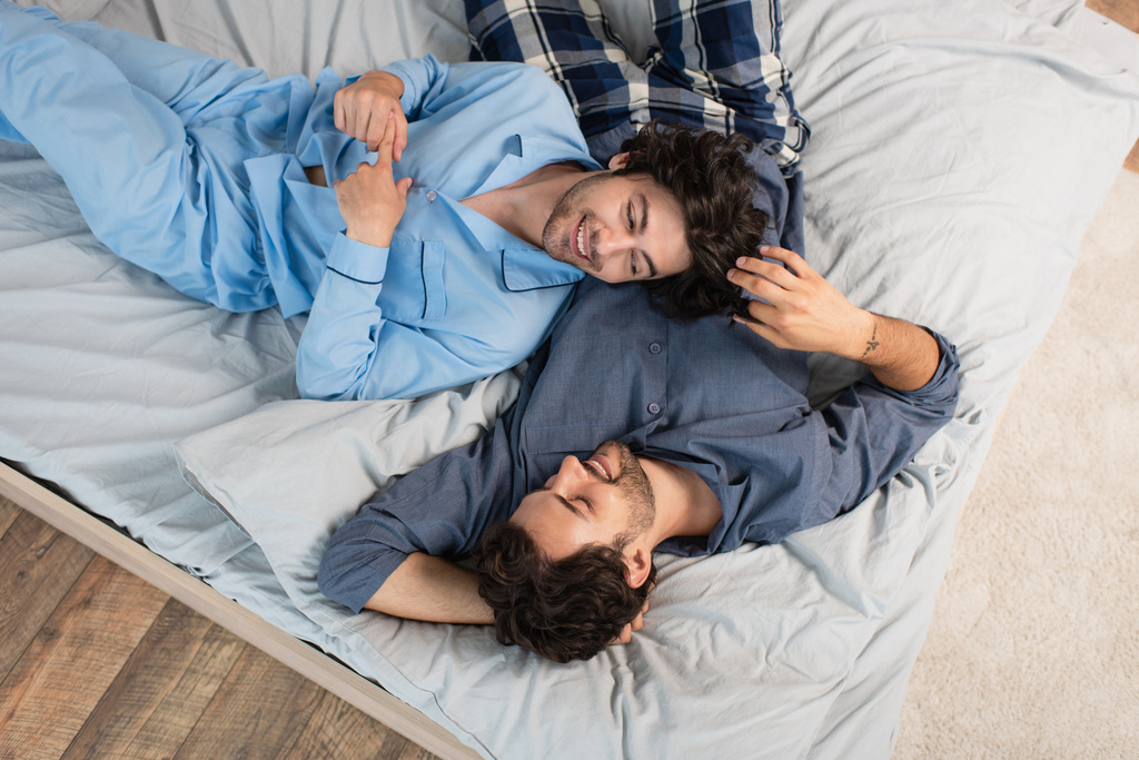 Blick von oben auf fröhliches homosexuelles Paar im Schlafanzug, das auf dem Bett ruht - Foto, Bild