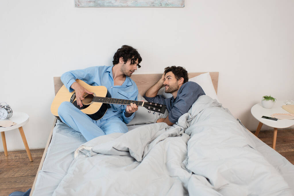 γκέι άντρας που παίζει ακουστική κιθάρα κοντά στο αγόρι του ξαπλωμένος στο κρεβάτι - Φωτογραφία, εικόνα