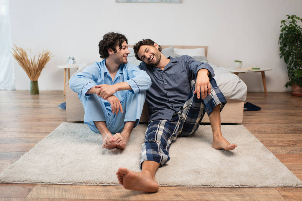 vrolijk jong gay paar zitten op tapijt in de buurt van bed - Foto, afbeelding