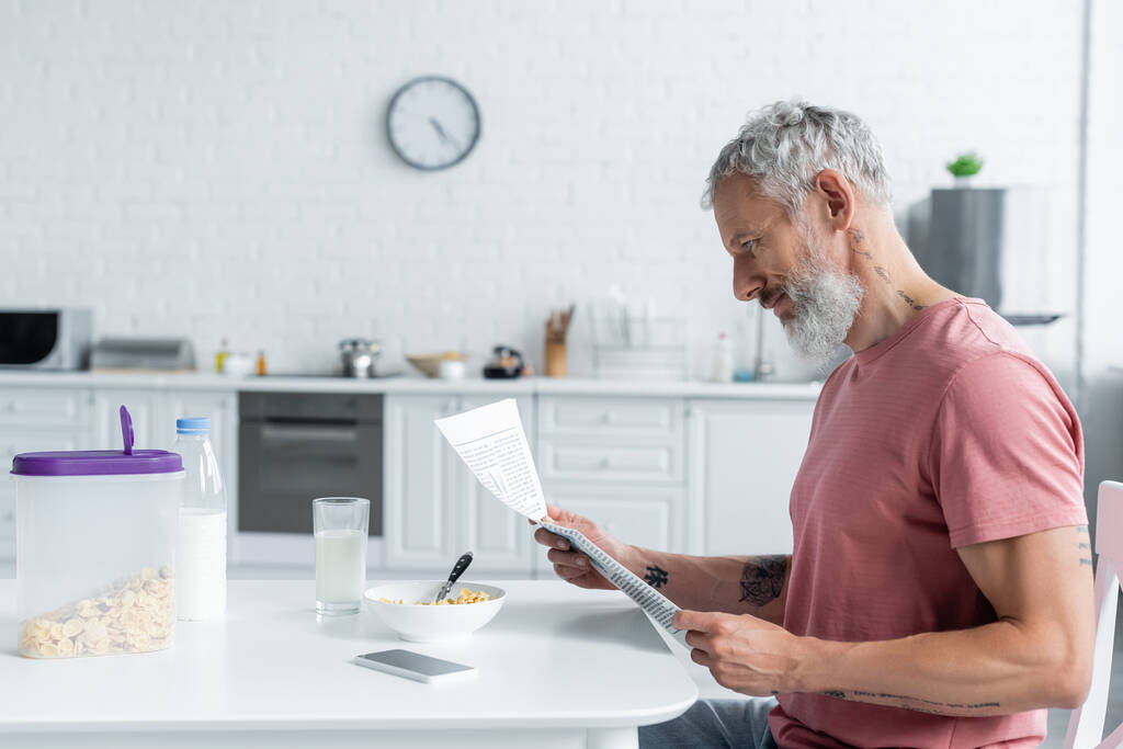 Side näkymä kypsä mies lukee uutisia lähellä älypuhelin ja aamiainen  - Valokuva, kuva
