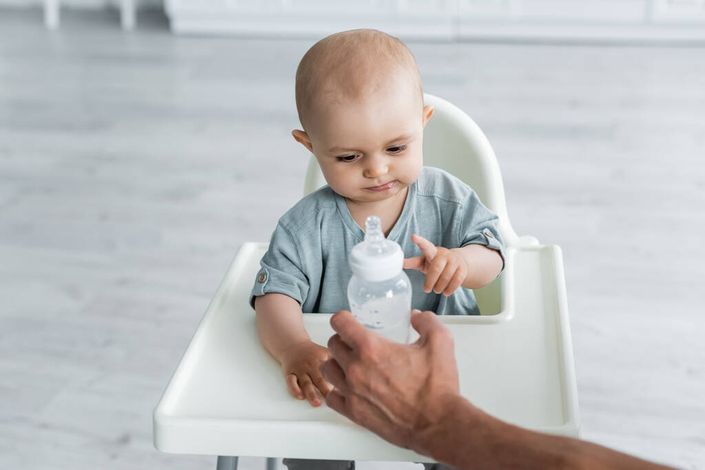Padre sosteniendo la botella con agua cerca del bebé en la silla alta  - Foto, Imagen