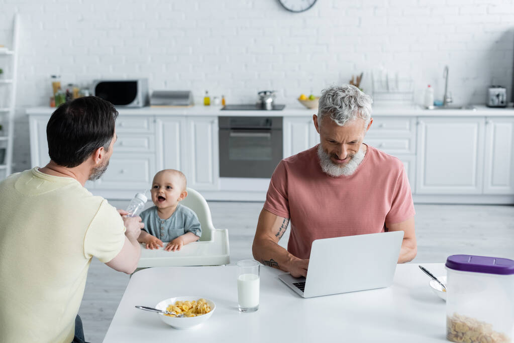 Homosexual hombre sosteniendo biberón cerca de bebé y pareja con portátil en la cocina  - Foto, imagen