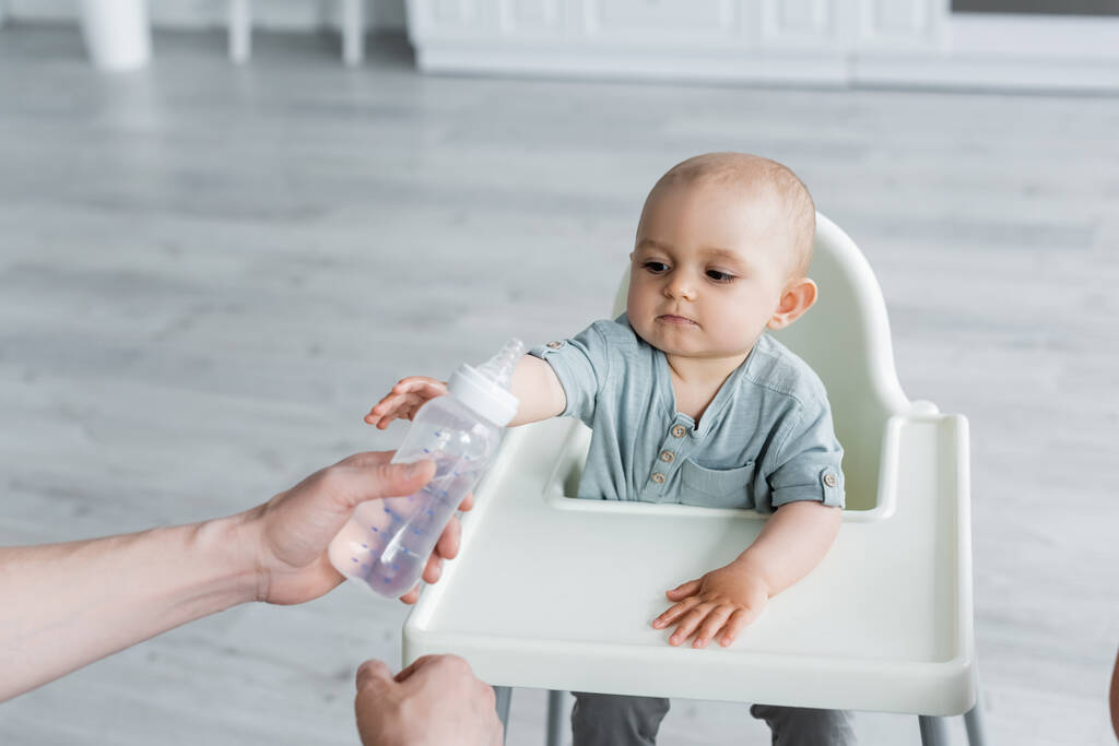 Otec drží kojeneckou láhev s vodou v blízkosti dítěte na vysoké židli  - Fotografie, Obrázek