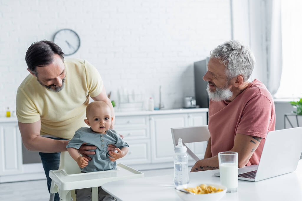 Smiling homosexual couple looking at baby daughter near laptop and breakfast  - Photo, Image