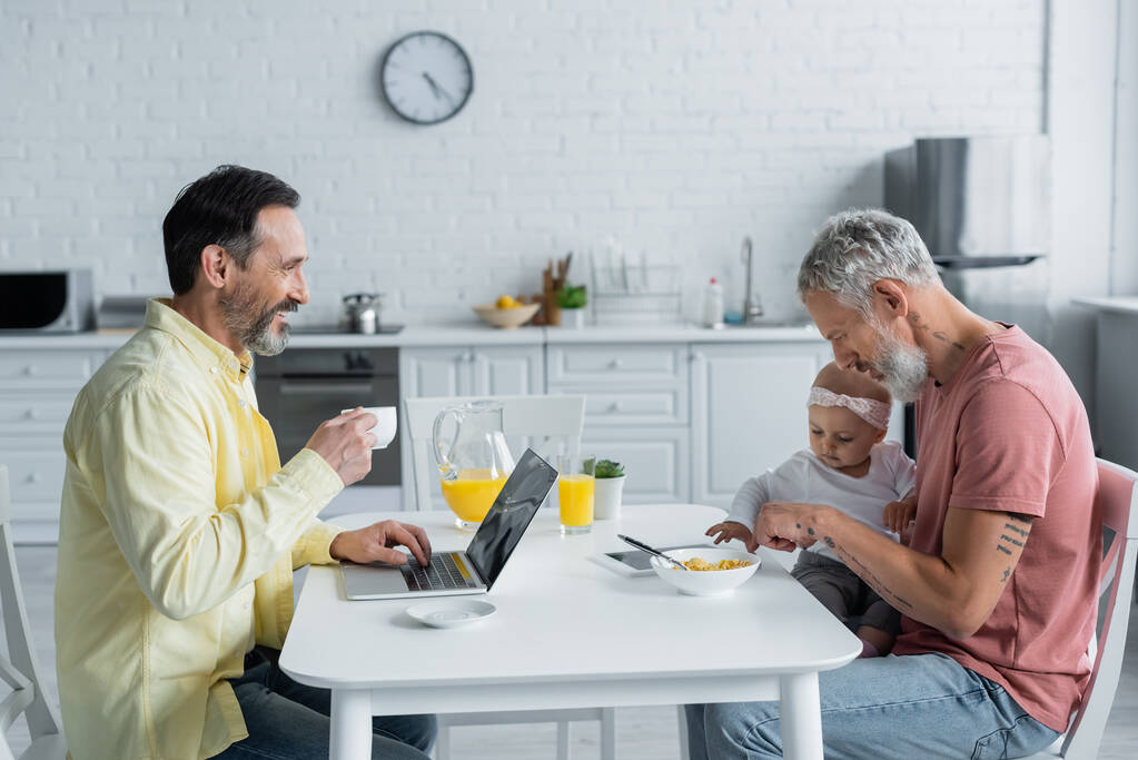 Χαμογελώντας ομοφυλόφιλος άνδρας με φορητό υπολογιστή και καφέ κοιτάζοντας σύντροφο με την κόρη στην κουζίνα  - Φωτογραφία, εικόνα