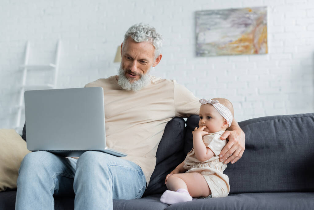 volwassen man holding laptop en omarmen baby meisje op bank  - Foto, afbeelding