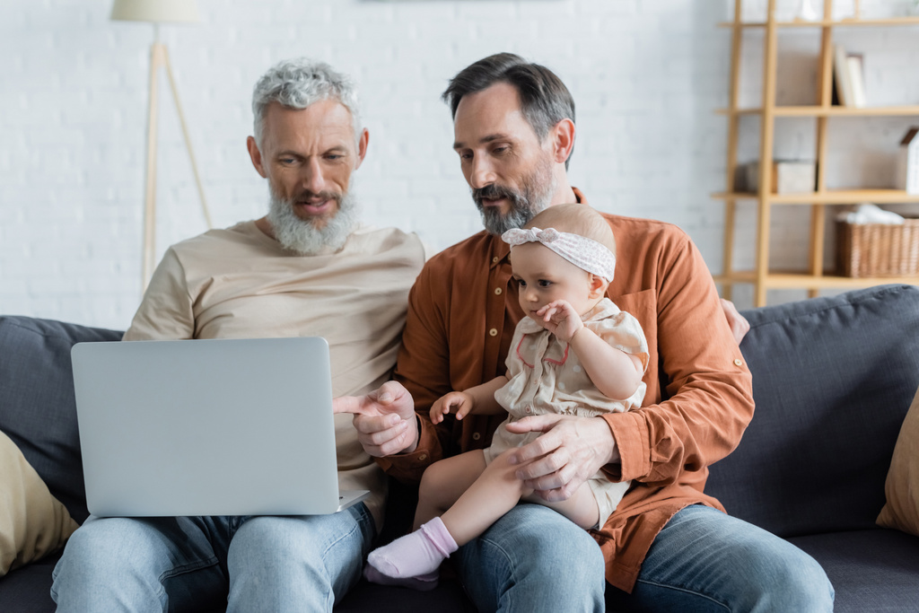Homosexuel homme pointant vers ordinateur portable près de bébé fille et partenaire à la maison  - Photo, image