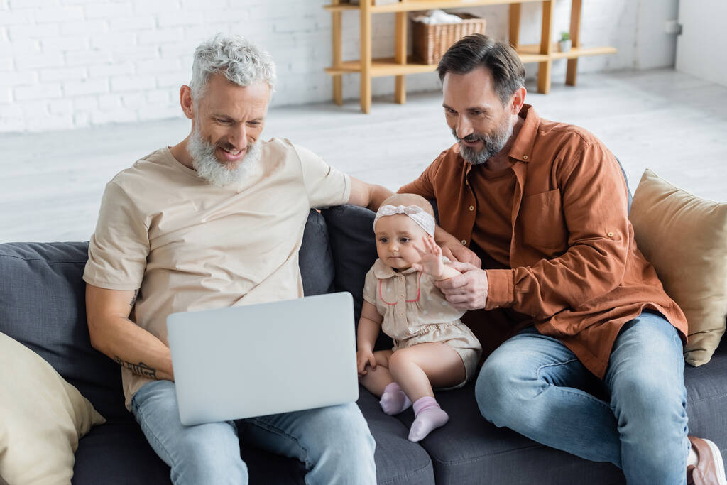Lächelndes gleichgeschlechtliches Paar nutzt Laptop neben Baby-Tochter auf Couch  - Foto, Bild