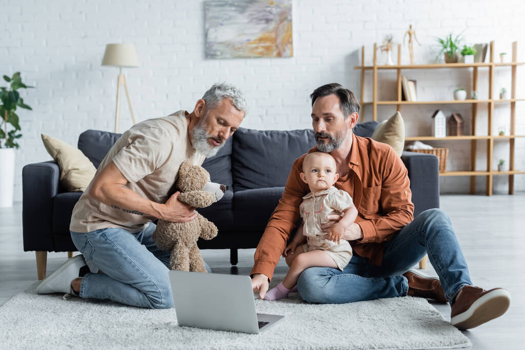 Familia del mismo sexo con la hija del bebé usando el ordenador portátil y sosteniendo juguete suave  - Foto, imagen