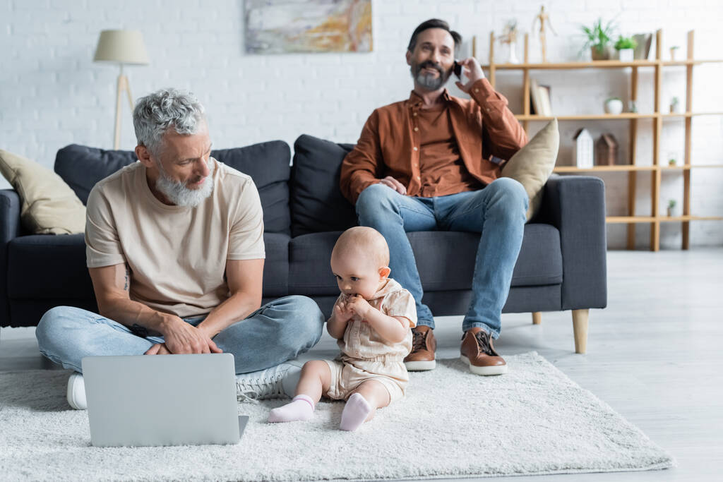 Dziecko dziecko siedzi w pobliżu homoseksualnego ojca z laptopem na dywanie  - Zdjęcie, obraz