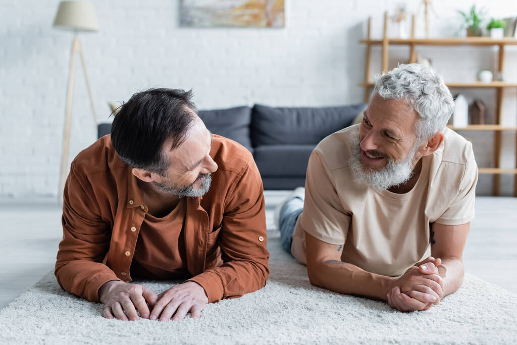 Cheerful homosexual couple looking at each other on carpet  - Photo, Image