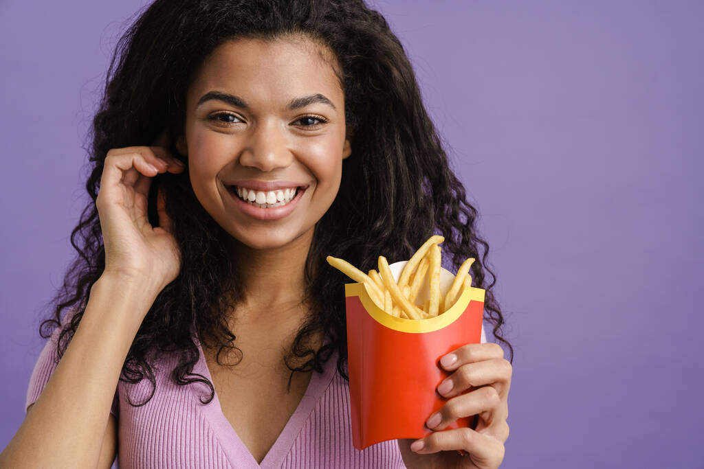 Joven mujer negra sonriendo mientras come papas fritas aisladas sobre fondo púrpura - Foto, Imagen