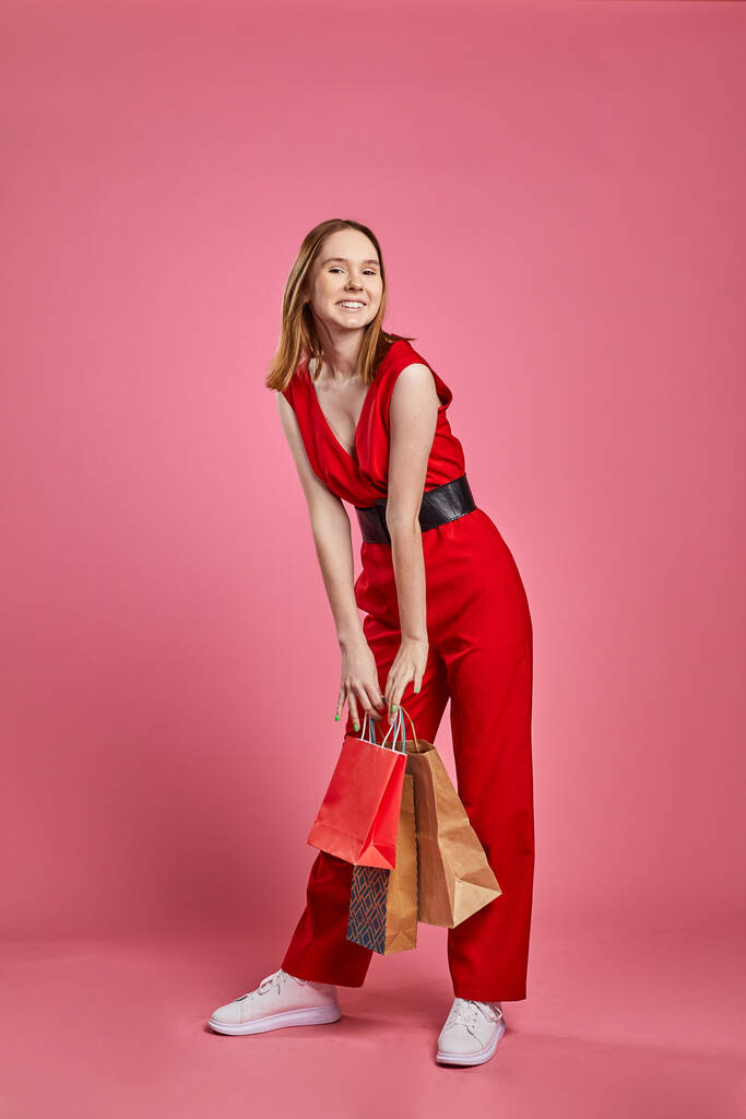 Fiatal nő boldog pozitív mosollyal visel elegáns piros ruhát izgatott hold bevásárló táskák eladó rózsaszín háttér. - Fotó, kép