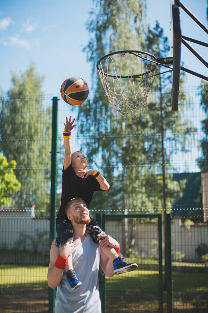 Glückliche Familie beim Basketball. Junger sportlicher Papa mit Sohn auf den Schultern. Junge wirft Ball in Basketballkorb - Foto, Bild