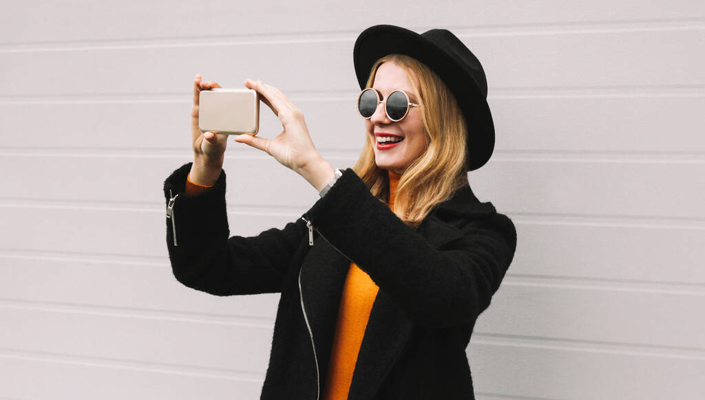 Портрет стильной веселой смеющейся молодой женщины, делающей селфи на смартфоне в черном пальто, круглой шляпе на сером фоне - Фото, изображение