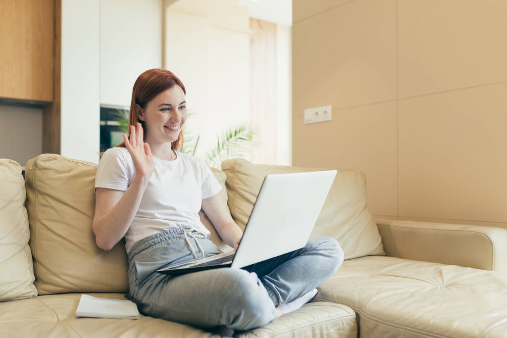 young happy girl talking on video call online with laptop webcam at home showing heart gesture. Caucasian red haired woman sitting on couch communicates remotely with a loved one Love sincere emotions - Photo, Image