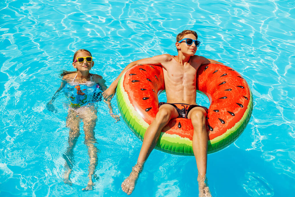 Happy kids, boy and girl in sunglasses, swim and play with inflatable rings, have fun and splash water in a beautiful pool in a tropical resort, have fun during family summer vacation - Photo, Image