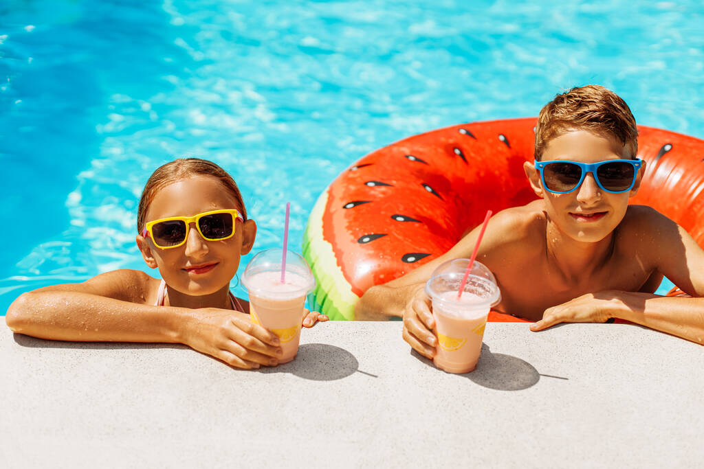 Zbliżenie wesoły mały chłopiec i dziewczyna w okularach przeciwsłonecznych picie koktajli podczas korzystania w basenie, Cute brat i siostra opierając się na krawędzi basenu i uśmiechnięty, Summer holiday concept - Zdjęcie, obraz