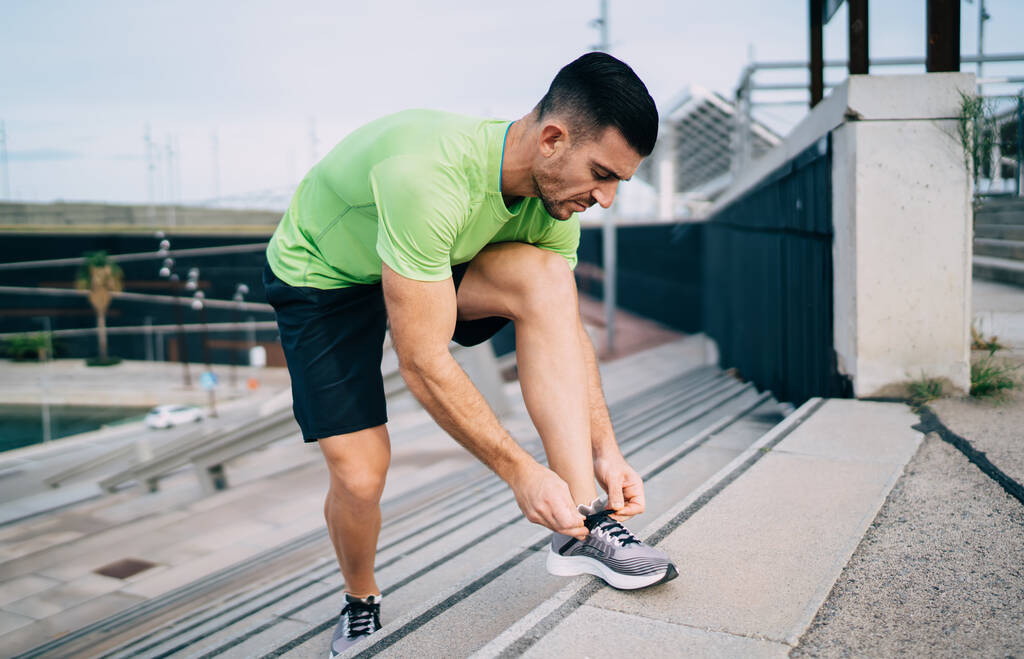 Valkoihoinen urheilija aktiivisissa vaatteissa ja trendikkäissä lenkkikengissä sitoo kengännauhat aamulla juoksentelemaan kaupungin kaduilla, päättäväinen mies lenkkeilijä 30s tarkistaa lenkkarit kardioharjoitteluun - Valokuva, kuva