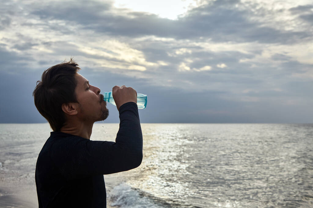 Чоловік п'є воду після пробіжки уздовж берега на тлі заходу сонця
 - Фото, зображення