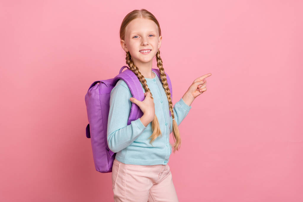 Photo of excited cute schoolgirl dressed blue cardigan smiling backpack pointing finger empty space isolated pink color background - Photo, Image