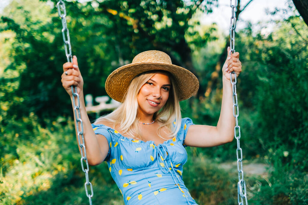 Portrait of attractive young woman in straw hat outdoors in summer sunny day on a swing. - Photo, Image