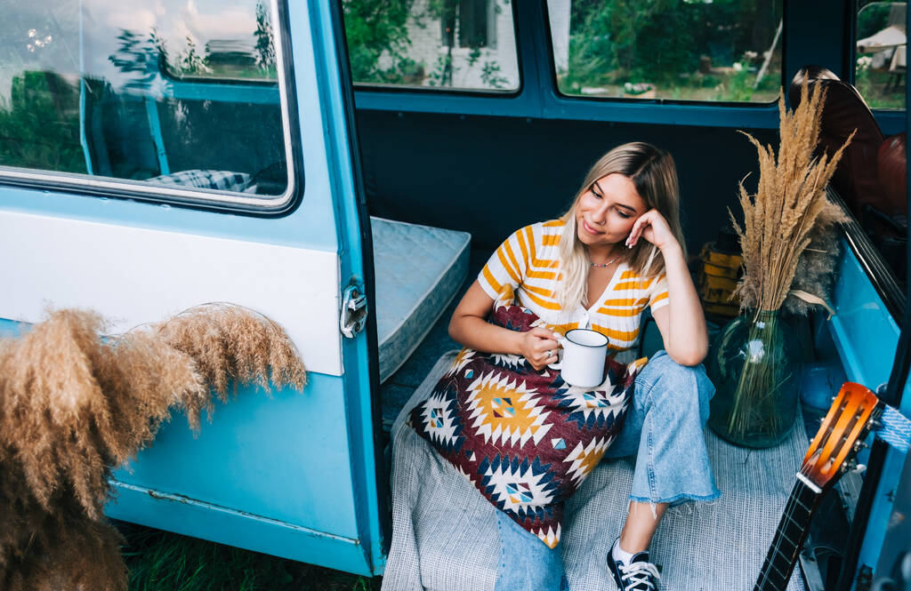 Retrato de uma jovem feliz sentada em uma van e bebendo chá ao ar livre na natureza, durante o pôr do sol. Desfrutando de verão, conceito de viagem - Foto, Imagem