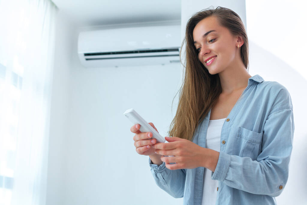 Young, happy, smile woman adjusts the temperature of the air conditioner using the remote control in room at home  - Photo, Image