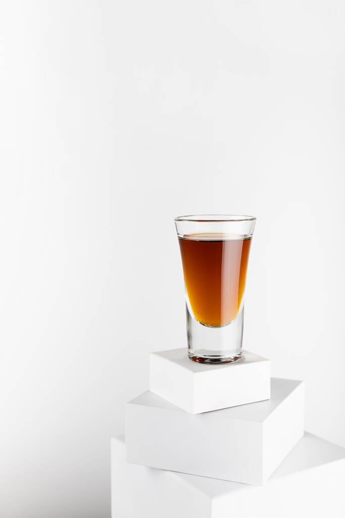 白い正方形の表彰台のスタックの上にショットガラスの古い黄金のコニャック、現代の最小限の幾何学的なスタイルで豪華なアルコール飲料、柔らかい光空間、垂直. - 写真・画像