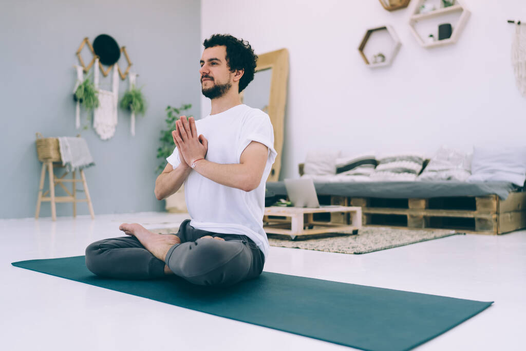 Sivunäkymä rauhallinen mies katselee pois istuu matto lootus poseeraa harjoitellessaan jooga viihtyisässä huoneessa koriste-elementtejä - Valokuva, kuva
